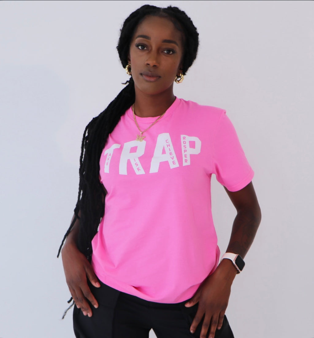 A person wearing a pink TRAP tee with a visible logo on the front with the words take risk achieve prosper standing against a white background